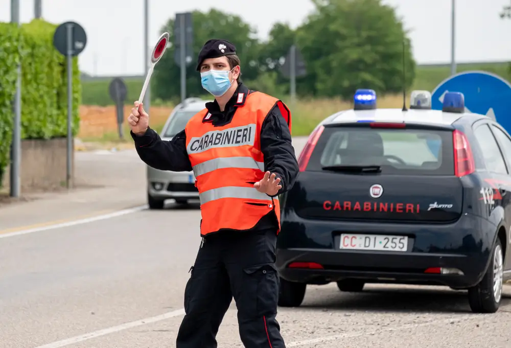 Carabiniere durante un controllo stradale con paletta segnaletica e auto di pattuglia con lampeggianti accesi.