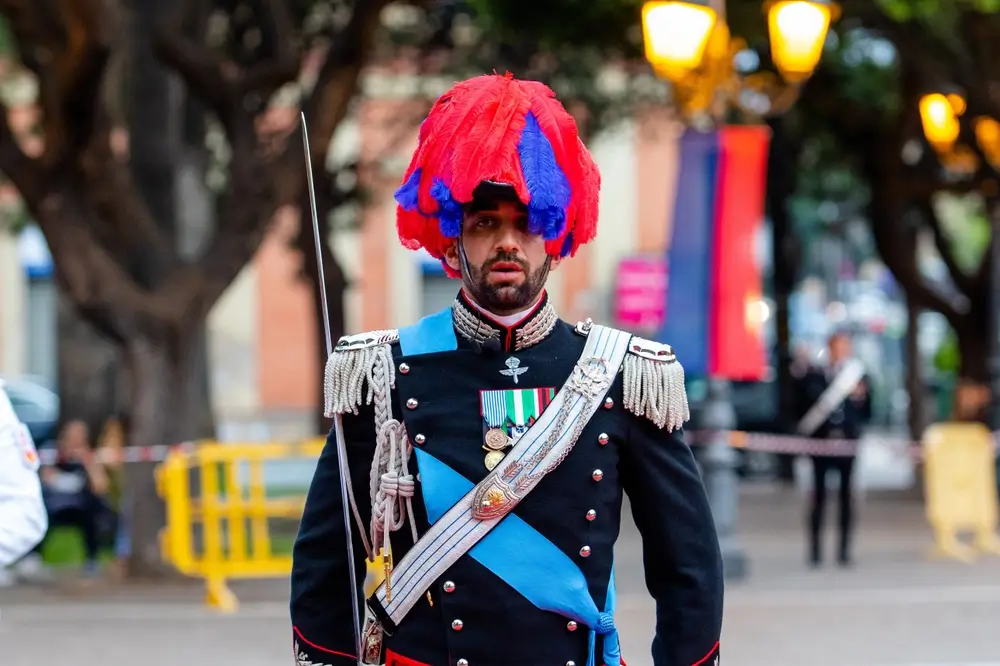 Ufficiale carabinieri in grande uniforme con pennacchio rosso e blu e sciarpa azzurra durante una cerimonia ufficiale.