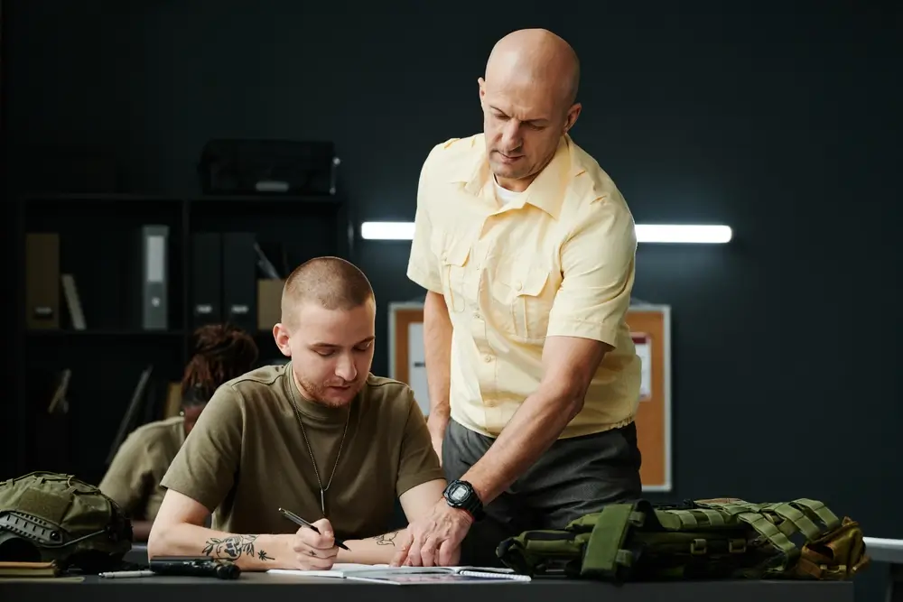 Allievo carabiniere scrive su un manuale durante il corso di formazione militare, assistito da un istruttore in aula.