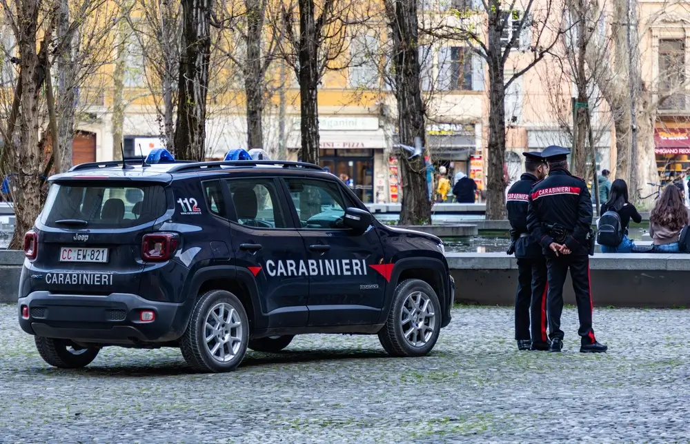 Pattuglia dei Carabinieri con brigadiere in servizio operativo in una piazza cittadina accanto all'auto d'ordinanza.