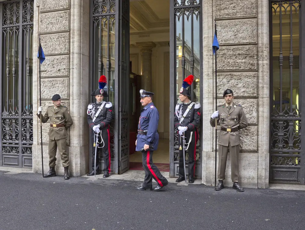 Maresciallo dei carabinieri attraversa l'ingresso di una stazione, affiancato da militari in uniforme cerimoniale.