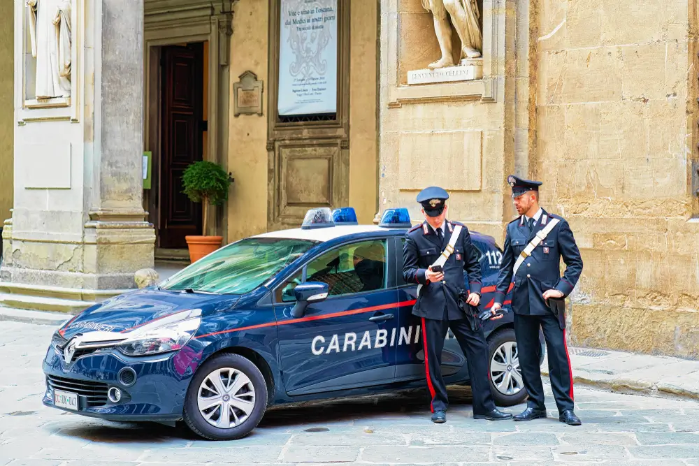 Due carabinieri in divisa accanto a un'auto di pattuglia blu in un centro storico cittadino.