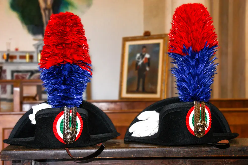 Due berretti della divisa dei Carabinieri con pennacchi rosso e blu e guanti bianchi su un banco di legno.