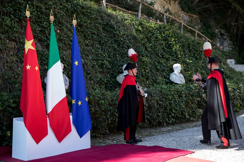 Maresciallo in divisa dei Carabinieri in alta uniforme con mantello e feluca accanto alle bandiere istituzionali.