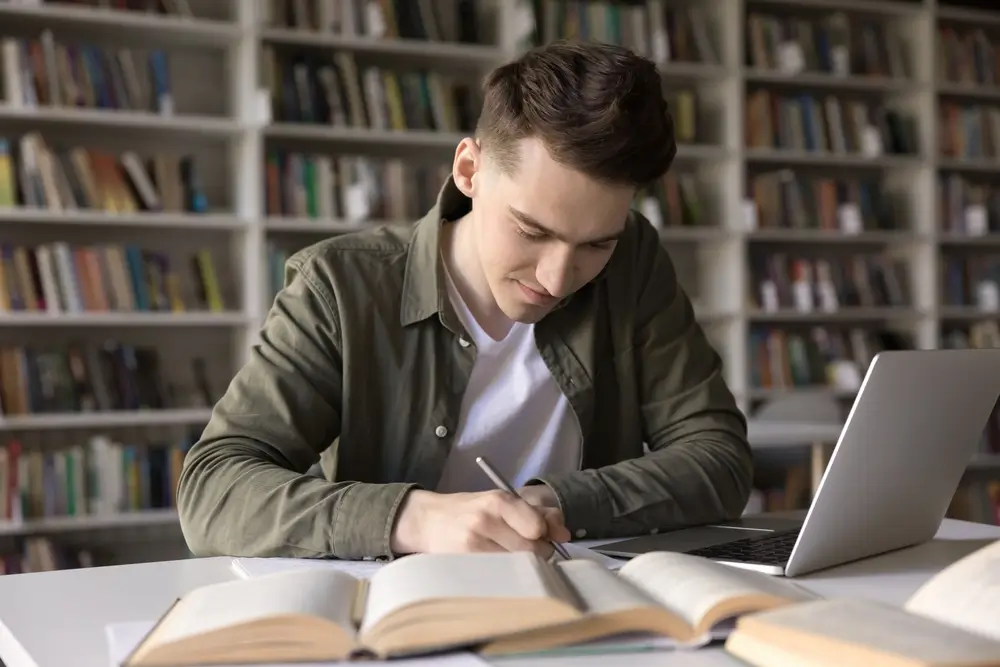 Candidato al concorso carabinieri studia in biblioteca con libri e laptop per la preparazione dell'esame.