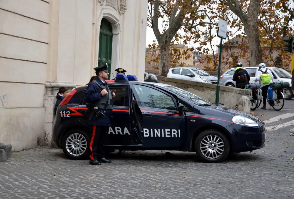 Carabiniere in divisa e cappello militare accanto a un'auto di pattuglia in una piazza cittadina.