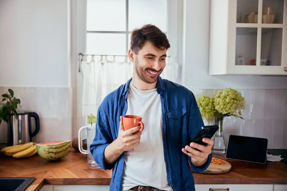 Giovane candidato sorridente consulta lo smartphone in cucina per verificare le notifiche ufficiali di un concorso.